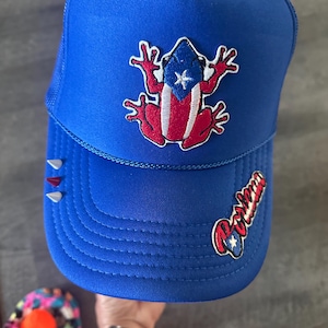 May include: A blue trucker hat with a red, white and blue frog with a Puerto Rican flag on its back. The hat has a white mesh back and the word "Boriken" embroidered on the side.