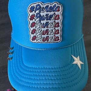 May include: A turquoise blue trucker hat with a white star on the side. The hat has a patch with the word "Purela" repeated five times in red, white, and blue.