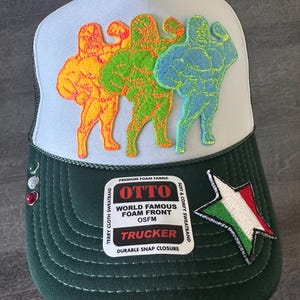 May include: A green and white trucker hat with a foam front. The hat features a patch with three cartoon muscle men in yellow, green, and blue. The patch also includes the text "OTTO WORLD FAMOUS FOAM FRONT OSFM TRUCKER DURABLE SNAP CLOSURE". The hat also has a small Italian flag star patch.
