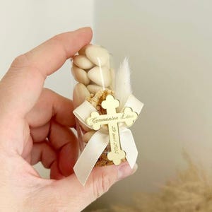 Puede incluir: Un tubo transparente lleno de caramelos blancos y una cruz decorativa dorada. La cruz tiene las palabras "Communion Léna" y "29 mai 2023" grabadas. Una cinta blanca y una pluma adornan el tubo.