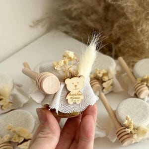 Pode incluir: Pequeno frasco de vidro cheio de mel, decorado com um tecido de juta e um amuleto de urso de madeira. O amuleto tem o texto "Baptême Romeo 11.06.23" gravado nele. Um mergulhador de mel de madeira está preso ao frasco.