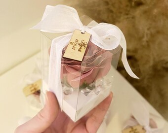 May include: A clear box with a pink rose inside. The box is tied with a white ribbon and a wooden tag that says "Ayma 05/01/23".