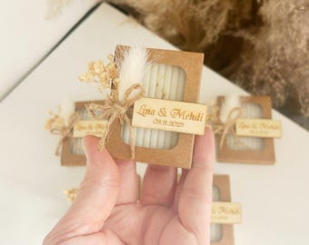 May include: Small, brown cardboard boxes with clear windows, each containing a white bar of soap. The boxes are decorated with dried flowers, twine, and a wooden tag with the text "Lina & Mehdi 08.11.2023".