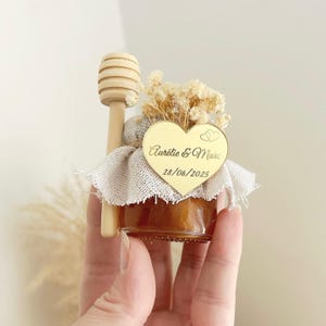 Peut inclure: Un petit pot de miel en verre, surmonté de fleurs séchées et d'un bâtonnet à miel en bois. Une étiquette dorée en forme de cœur porte les noms "Aurélie & Marc" et la date "28/06/2025". Le pot est enveloppé dans un tissu blanc.