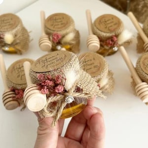 Peut inclure: Petits pots de miel en verre, décorés de jute, de couvercles en bois et de fleurs séchées. Chaque pot est accompagné d'un bâtonnet à miel en bois. Les couvercles ont du texte imprimé.