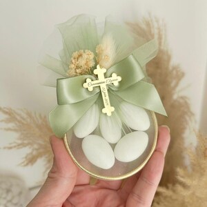 Puede incluir: Una pequeña caja de regalo redonda con borde dorado, llena de caramelos blancos. La caja está adornada con una cinta verde salvia, una cruz dorada y una tela verde transparente. La cruz está grabada con texto. También se incluyen flores secas y trigo.