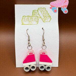 May include: A pair of handmade earrings featuring a bright pink and white design. The earrings are made from plastic blocks and have silver-colored hooks. The image also includes a pink roller skate illustration and green block drawings.