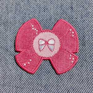 Puede incluir: Un parche bordado con forma de lazo rosa con un centro blanco que presenta un diseño de lazo más pequeño. El parche está sobre un fondo de mezclilla azul. El lazo tiene un borde decorativo cosido.