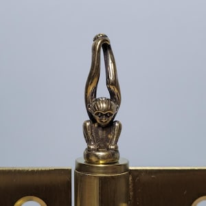 May include: Brass hinge featuring a decorative monkey figure. The hinge has two rectangular mounting plates and a central barrel. The monkey figure is seated with its arms raised above its head.