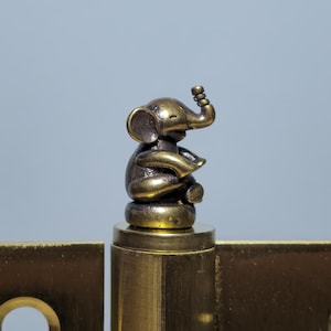 Puede incluir: Bisagra de latón con una figura decorativa de elefante. La bisagra tiene dos placas rectangulares con agujeros circulares. El elefante está detallado con una trompa y orejas.