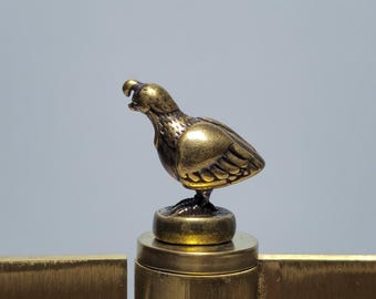 May include: A brass hinge with a decorative bird figurine on top. The hinge has a rectangular shape with two circular holes. The bird is detailed with feathers and a beak.