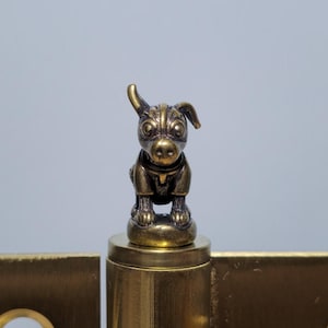 May include: A brass-coloured hinge featuring a small, detailed dog figurine. The hinge has a rectangular shape with rounded edges and circular holes. The dog is in a sitting position.