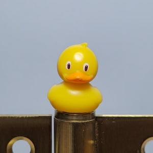 Könnte beinhalten: Ein Messing-Scharnier mit einer gelben Quietscheente darauf. Das Scharnier hat eine rechteckige Form mit drei kreisförmigen Löchern. Die Ente hat einen gelben Körper, einen orangefarbenen Schnabel und schwarze Augen.