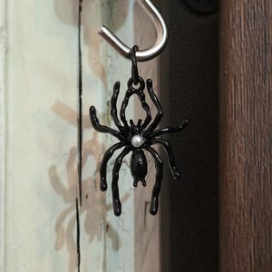 May include: A black spider charm with a pearl-like accent hangs from a silver hook. The spider has eight legs and a detailed body. The charm is suspended against a backdrop of a wooden door and a painted wall.