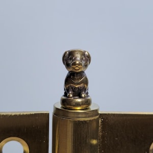 Puede incluir: Una bisagra de latón con una figura decorativa de perro. La bisagra es de color dorado brillante, y el perro está sentado. La bisagra tiene dos agujeros circulares y está sobre un fondo gris claro.