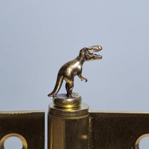 May include: A brass hinge with a small, detailed Tyrannosaurus Rex figurine on top. The hinge is a warm gold color, and the dinosaur is a darker, antique gold. The background is a soft, muted gray.