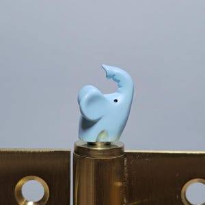 Peut inclure: Une charnière en laiton avec une figurine d'éléphant bleu clair. L'éléphant a sa trompe levée et est monté sur un cylindre en laiton. La charnière est rectangulaire avec deux trous circulaires.