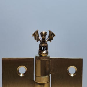 Op de afbeelding: Een messing deur scharnier met een klein drakenbeeldje erop. De draak zit op een ronde basis en heeft zijn vleugels uitgespreid.