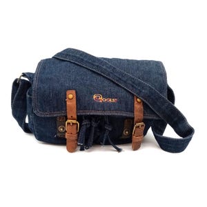 Puede incluir: Un bolso bandolera de mezclilla azul con correas y hebillas de cuero marrón. El bolso tiene un detalle de borla y la palabra "COOLST" bordada en la parte delantera.