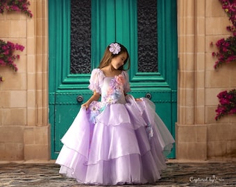 May include: A young girl wearing a lavender dress with floral embellishments stands in front of a teal double door. The dress has multiple layers of tulle and a floral design on the bodice. The girl is smiling and looking down at the dress.