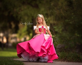 May include: A young girl wearing a pink and red princess dress with a gold crown. The dress has a layered skirt and white accents. The girl is standing on a paved path in a park-like setting with green foliage in the background.