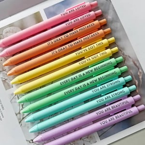 Może przedstawiać: Kolekcja pastelowych długopisów ułożonych w gradient tęczy. Każdy długopis ma inny inspirujący napis, np. "You Are Loved", "It's Okay to Make Mistakes" i "You've Got This". Idealne do pisania.