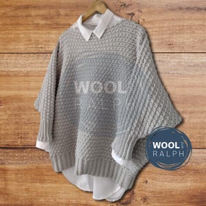 Op de afbeelding: Een grijze gebreide trui met een losse pasvorm en een brede, lage halslijn. De trui heeft een textuurpatroon en hangt aan een houten hanger. De merknaam "WOOL and RALPH" is zichtbaar op de trui.