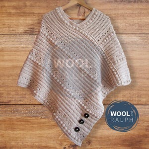 Peut inclure: Un poncho en crochet beige avec un col en V et un motif en diagonale. Le poncho a un design texturé avec de petites pompons blanches et trois boutons noirs. Le poncho est suspendu à un cintre en bois.