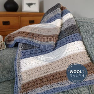 Könnte beinhalten: Eine handgefertigte gehäkelte Decke mit Streifen in Blau-, Weiß-, Grau- und Brauntönen. Die Decke ist über einem Sofa drapiert und zeigt ihre Textur und ihr Design. Die Decke hat einen blauen Rand und den Text "WOOL and RALPH".