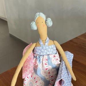 May include: A handmade doll with light blue yarn hair, wearing a blue and pink patterned dress and holding a pink and blue patterned blanket.