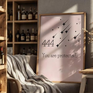 Könnte beinhalten: Ein gerahmter Druck mit rosa Hintergrund zeigt die Zahl "444" und den Text "You are protected." Schwarze Sternschnuppen-Grafiken sind ebenfalls zu sehen. Der Druck wird in einem Raum mit einem Holzregal und einem gemütlichen Stuhl ausgestellt.