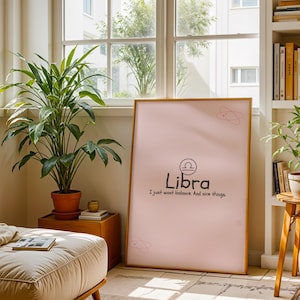 May include: A framed pink Libra zodiac sign art print. The print features the Libra symbol and the words "Libra, I just want balance. And nice things." The artwork is displayed in a well-lit room with plants and a window.
