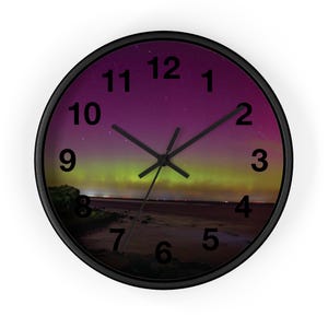 Pode incluir: Um relógio de parede redondo com moldura preta. O mostrador do relógio apresenta uma imagem panorâmica da aurora boreal com um céu roxo e verde sobre uma paisagem escura. Ponteiros e algarismos pretos completam o design.