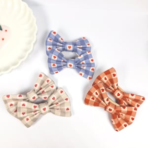 May include: Three pairs of fabric hair bows with a gingham pattern and red heart accents. The bows are in blue, beige, and orange.