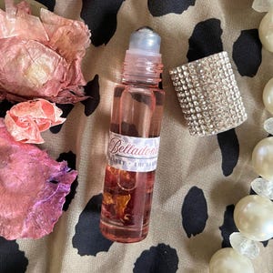 May include: A clear glass rollerball bottle filled with a pink liquid and visible dried flower petals. The bottle features a white label with the text Belladonna Beauty Enchantment and a separate cap covered in small, clear, reflective rhinestones.