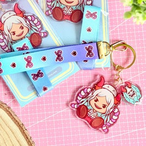 Könnte beinhalten: Acryl-Schlüsselanhänger und Lanyard-Set mit einer Zeichentrickfigur mit weißen Haaren, roten Hörnern und einem rosa-weißen Outfit. Das Lanyard ist blau mit einem sich wiederholenden Muster aus Schleifen und Diamanten. Der Schlüsselanhänger hat einen goldenen Verschluss.
