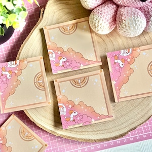 Può includere: Biglietti adesivi quadrati beige con un design a nuvola rosa e arancione, una rosa dei venti e un personaggio dei cartoni animati. I biglietti sono disposti su una fetta di legno, con un polpo all'uncinetto rosa e bianco sullo sfondo.