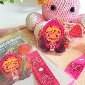 May include: Acrylic keychain with a cartoon character in a pink hooded jacket and blonde hair. The keychain includes a heart-shaped charm and a pink lanyard. The packaging reads "Ori's merch studio Lanyard Acrylic Keychain."