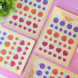 Op de afbeelding: Vier stickervellen met illustraties van harten en fruit in rood, oranje en paars. De stickers zijn gerangschikt op rechthoekige vellen. De tekst "Ori's Merch Studio" is afgedrukt.