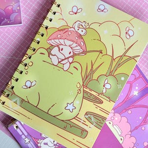 Könnte beinhalten: Ein spiralgebundenes Notizbuch mit einer skurrilen Illustration. Das Cover zeigt eine Cartoon-Frosch- und Pilzszene in Grün-, Gelb- und Rosatönen. Schmetterlinge und Sterne sind um das Design herum verstreut. Das Notizbuch liegt auf einer rosa Schneidematte.