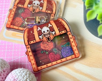 May include: A clear acrylic keychain shaped like a treasure chest with a skull and crossbones design. The chest is filled with three colorful cartoon fruits. The keychain has a gold metal ring.