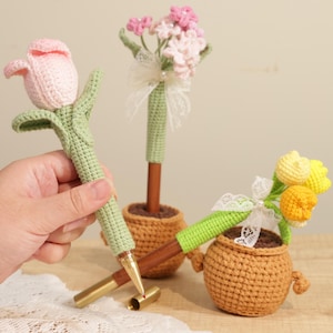 May include: Crocheted flower pens in pink, yellow, and orange, with green stems and leaves. The pens are in small brown crocheted pots. One pen is held in a hand. The pens have gold-colored tips and wooden barrels.