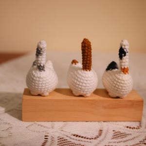 May include: Three handmade crochet cat butt ornaments in white with gray, brown, and black tail details. The cat butts are displayed on a small, rectangular wooden block, perfect for cat lovers and home decor.