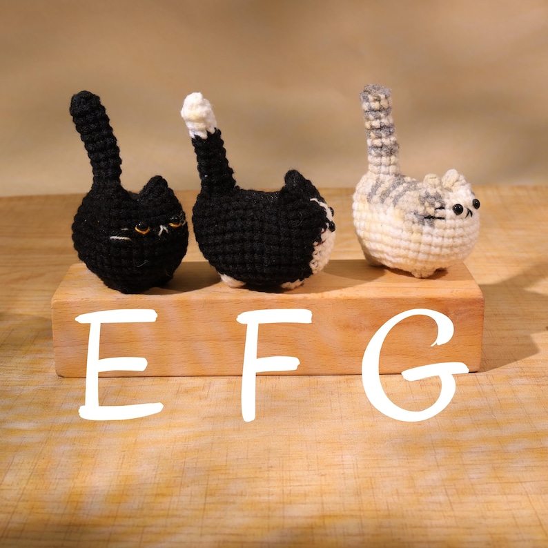 May include: Three crocheted cat figurines in black, black and white, and gray and white. The cats are sitting on a wooden block, with the letters "EFG" in white below them. The cats have upright tails and small, detailed faces.