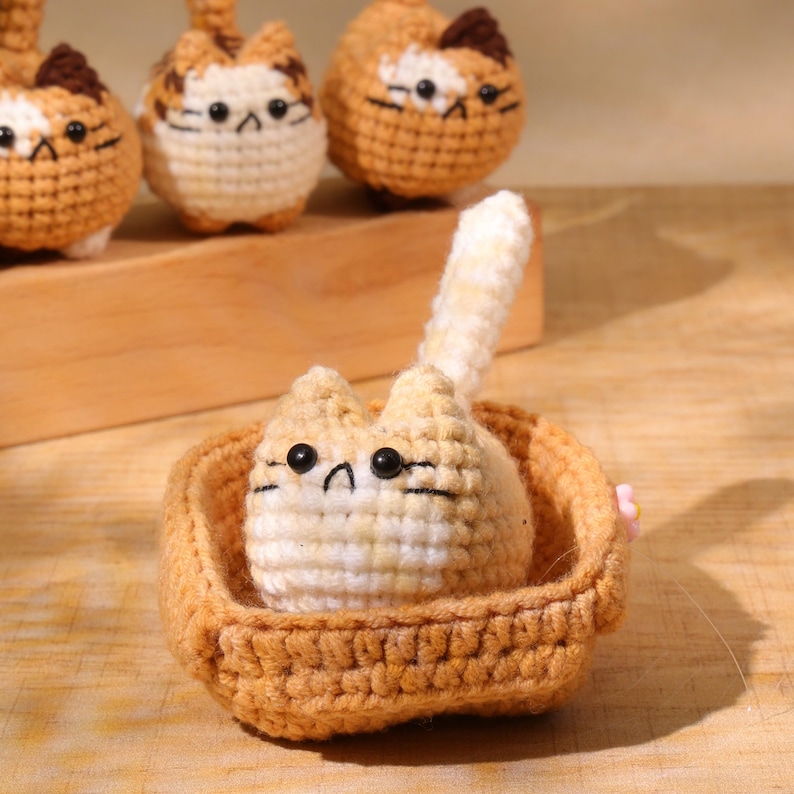 May include: Crocheted cat figurine in a woven basket. The cat is tan and white with black eyes and whiskers. The basket is a light brown color. Several other cat figurines are in the background.