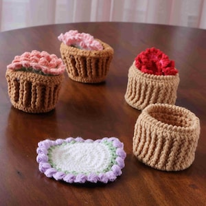 May include: A selection of handmade crochet cupcake-shaped containers and a coaster. The cupcakes have brown bases and colourful, textured tops in pink and red. The coaster is white with a lavender and green border. The items are on a wooden surface.