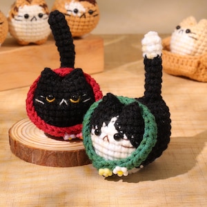 May include: Crocheted cat figurines in black and white, and black with red trim. The cat figures have yellow eyes and small white flowers at the base. Other cat figures are visible in the background.