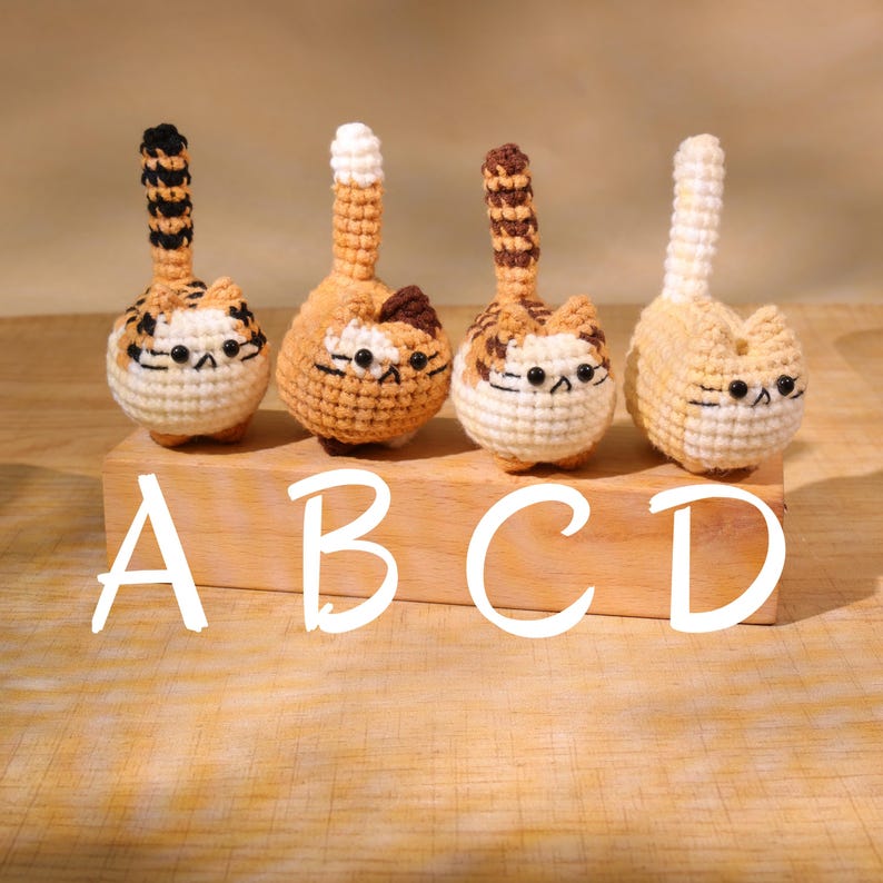 May include: Four handmade crochet cat figurines in cream, orange, and brown. Each cat has a striped tail and a detailed face. The figurines are displayed on a wooden block with the letters A, B, C, and D in white.