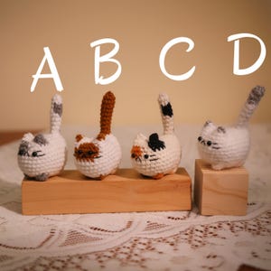 May include: Four handmade crochet cat figurines in white, with gray, brown, and orange markings. Each cat has a tall tail and is displayed on a wooden base. Letters A, B, C, and D are above the cats.
