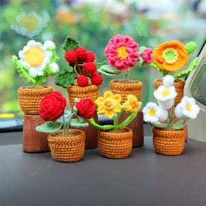 May include: A collection of handmade crochet flowers in small brown pots. The flowers are various colors, including white, red, pink, yellow, and orange. The pots are arranged on a dark surface, with a blurred green background.